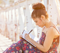 Image of a woman writing in a journal while sitting on a small bridge
